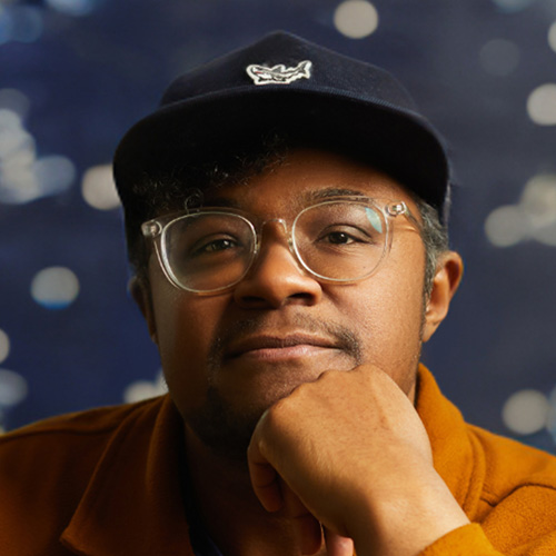 Headshot of man with warm brown skintone and brown eyes with glasses and hat