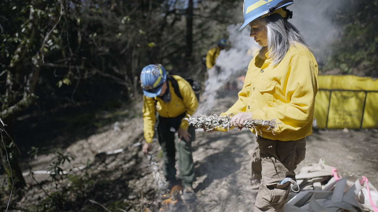 Women in protective gear standing near wild fire holding some plants
