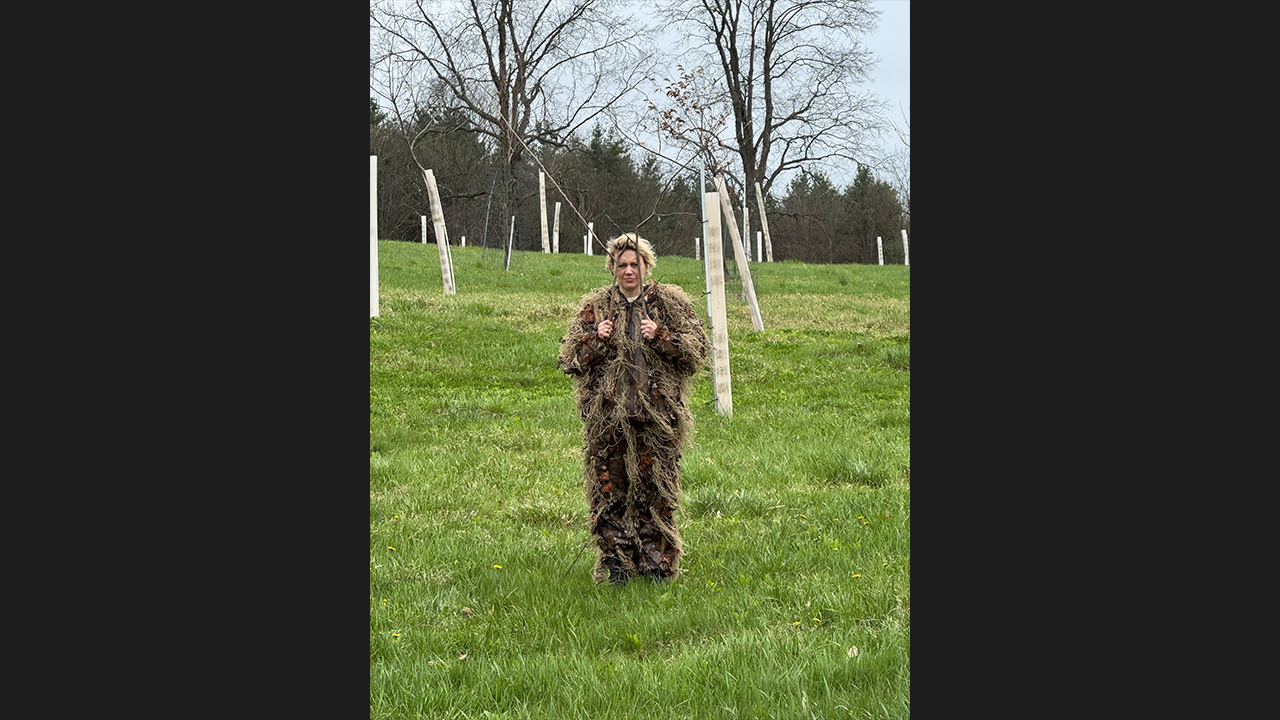 Kate McKinnon standing in a ghillie suit holding sticks up to her face