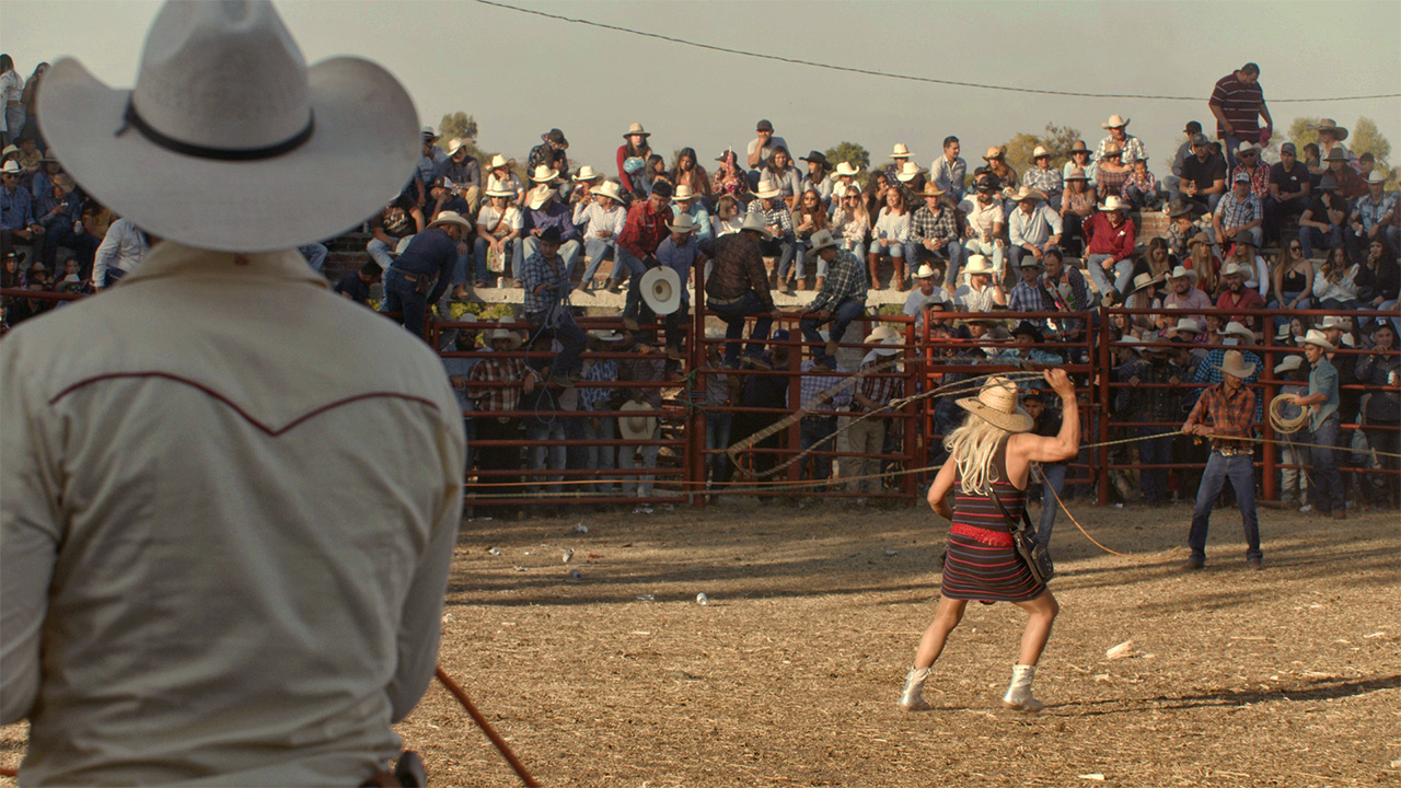 Person in dress and blond wig lassoing animal off frame with cowboys all around