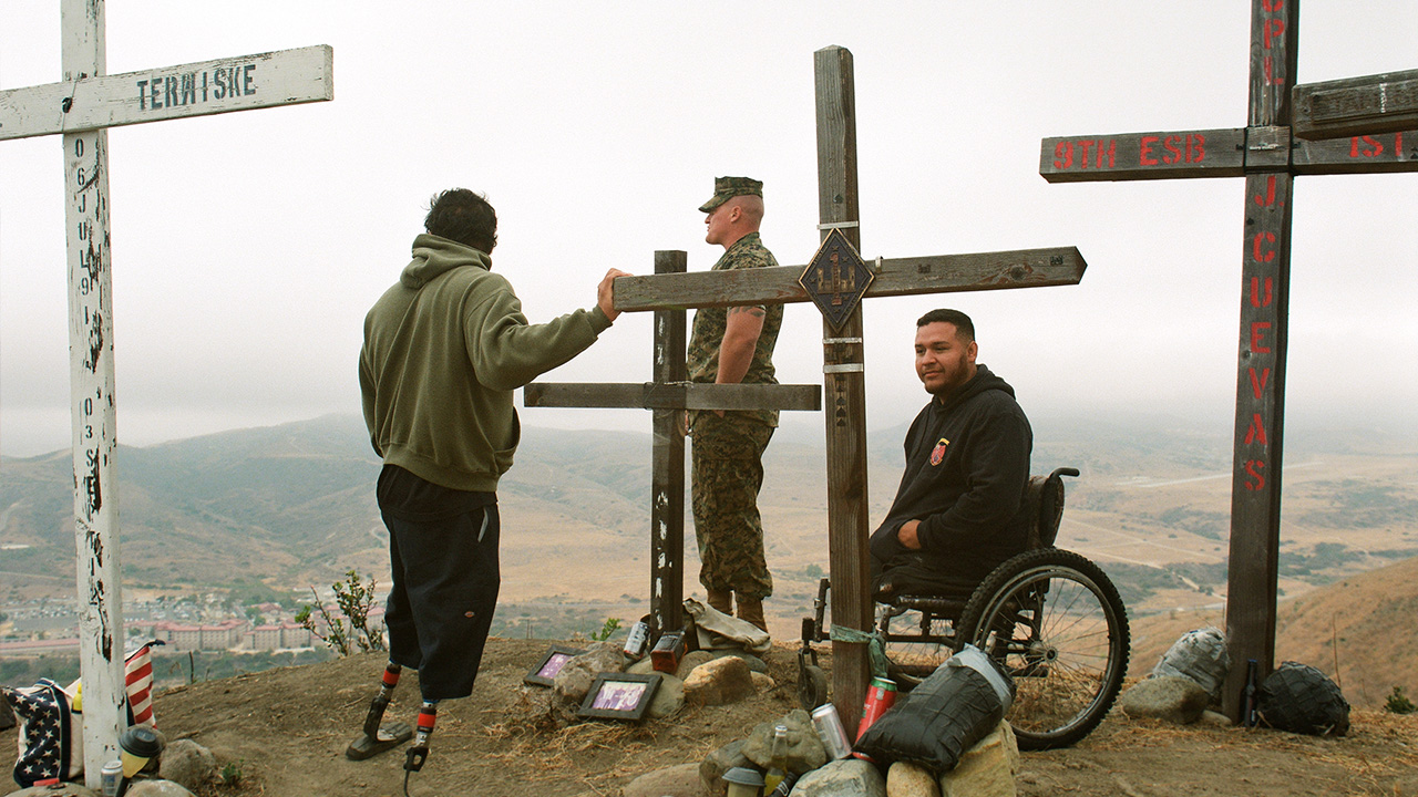 Three men one in army uniform and one in wheelchair standing along tall crosses in the ground that look like memorials