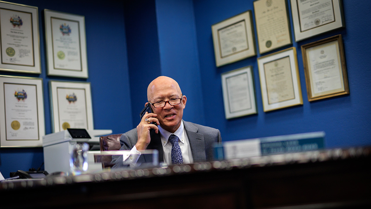 man sitting at desk with lots of plaques on the wall talking on the phone