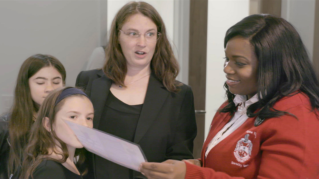 Woman wearing blazer with crest on it and dark skin tone talkinhg with a woman and her two young daughters who are also dressed up. Everyone looking at a piece of paper