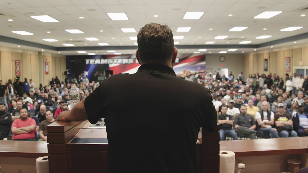 Man talking to a large group from a stage with a large poster that says 'teamsters' in the back of the room