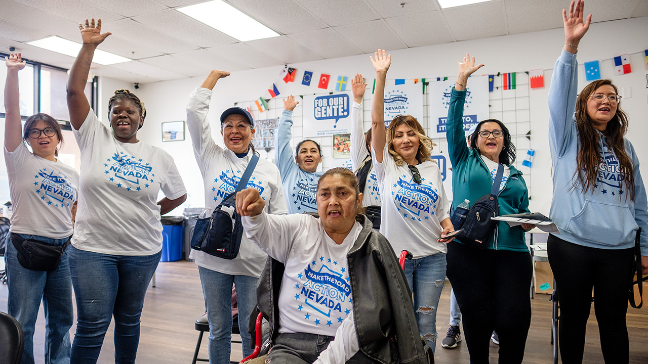 a group of women of color holding their hands in the air with matching T-shirts that say 'Make The Road. Action Nevada'