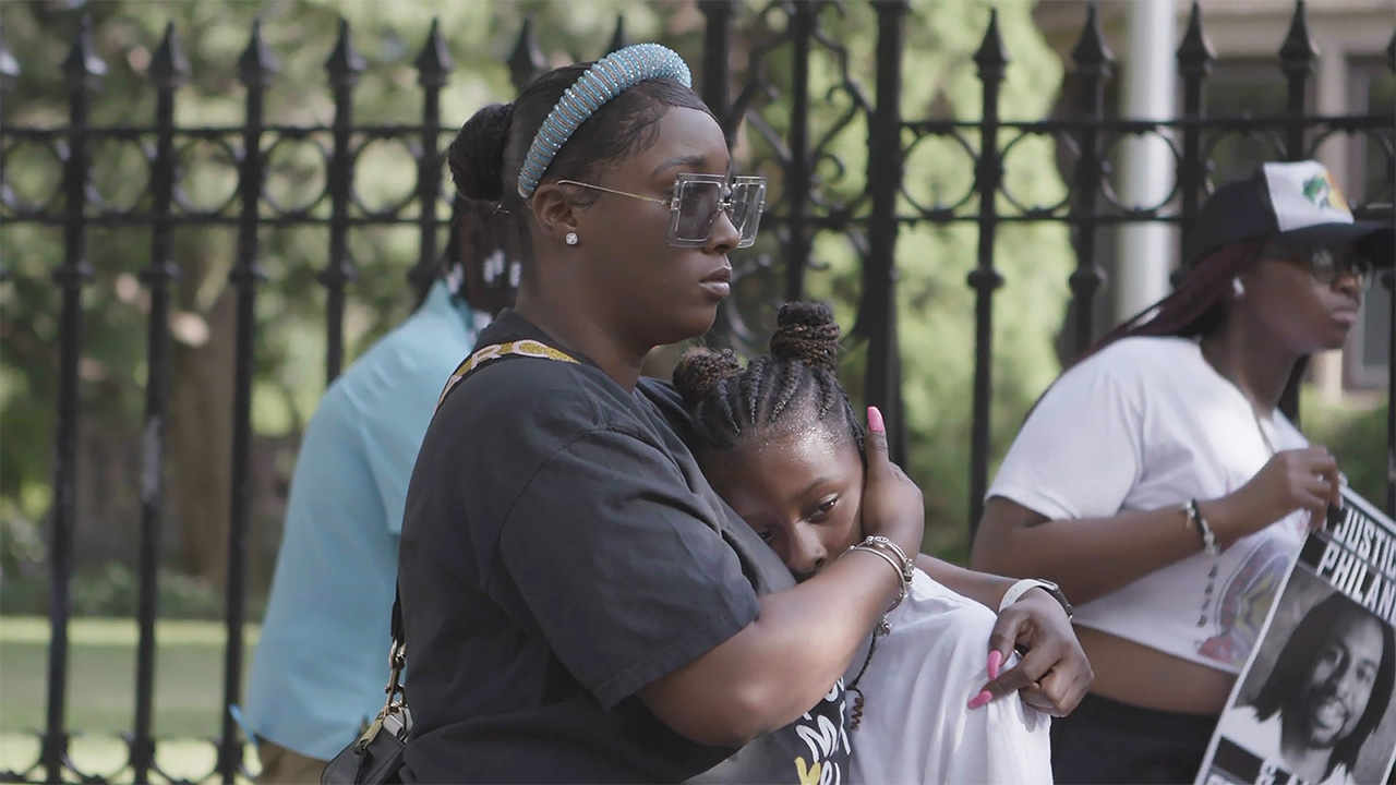 Woman with dark brown skin tone standing comforting a child while they look off the side of the image