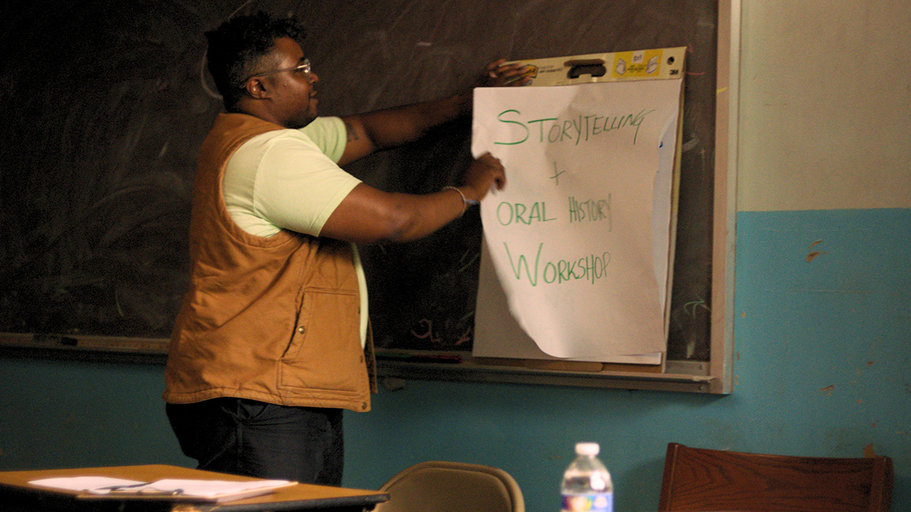 Man standing by chalk board ripping paper off xl stickypad that says "storytelling and Oral History workshop"