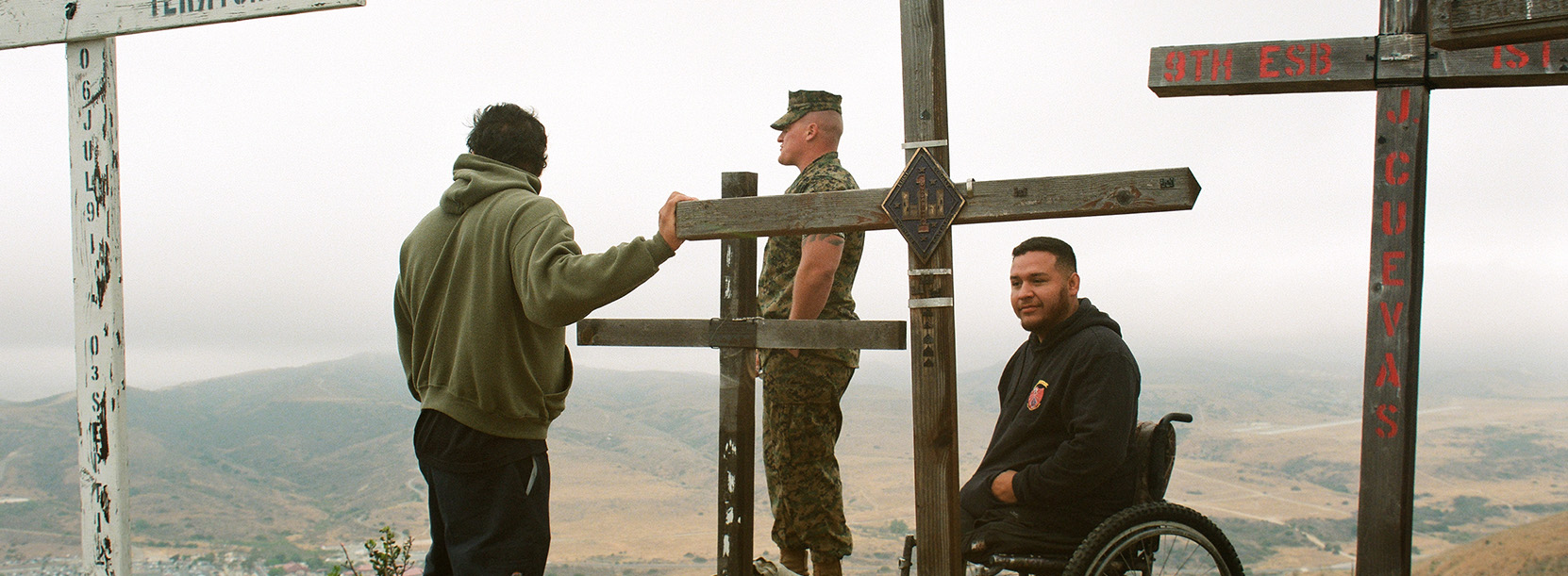 three men, one in military uniform, one in wheel chair and another in regular clothes standing next to wooden crosses on a hill top